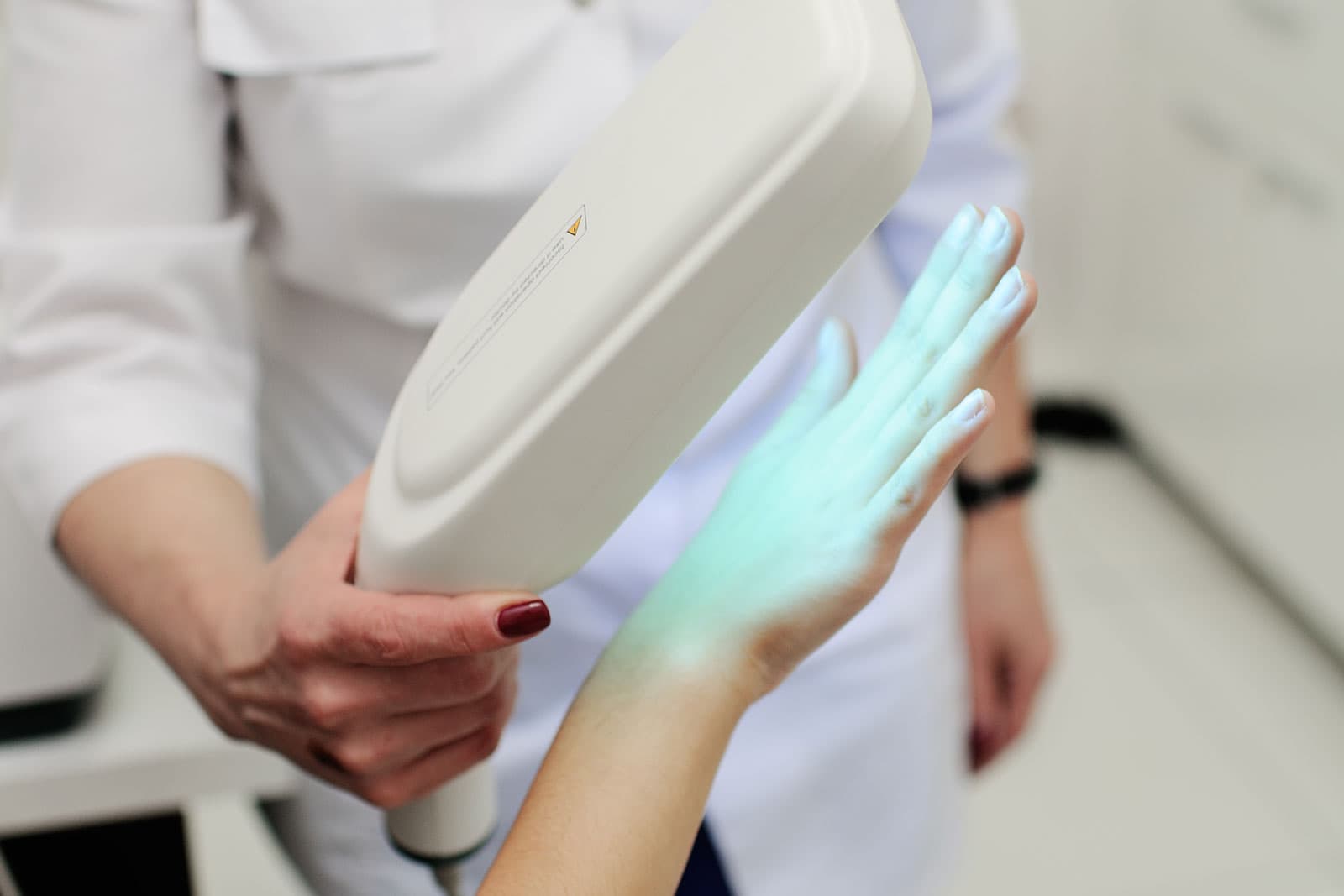 Clinician treating a patient's hand with a phototherapy device emitting blue-green light during a skin treatment.